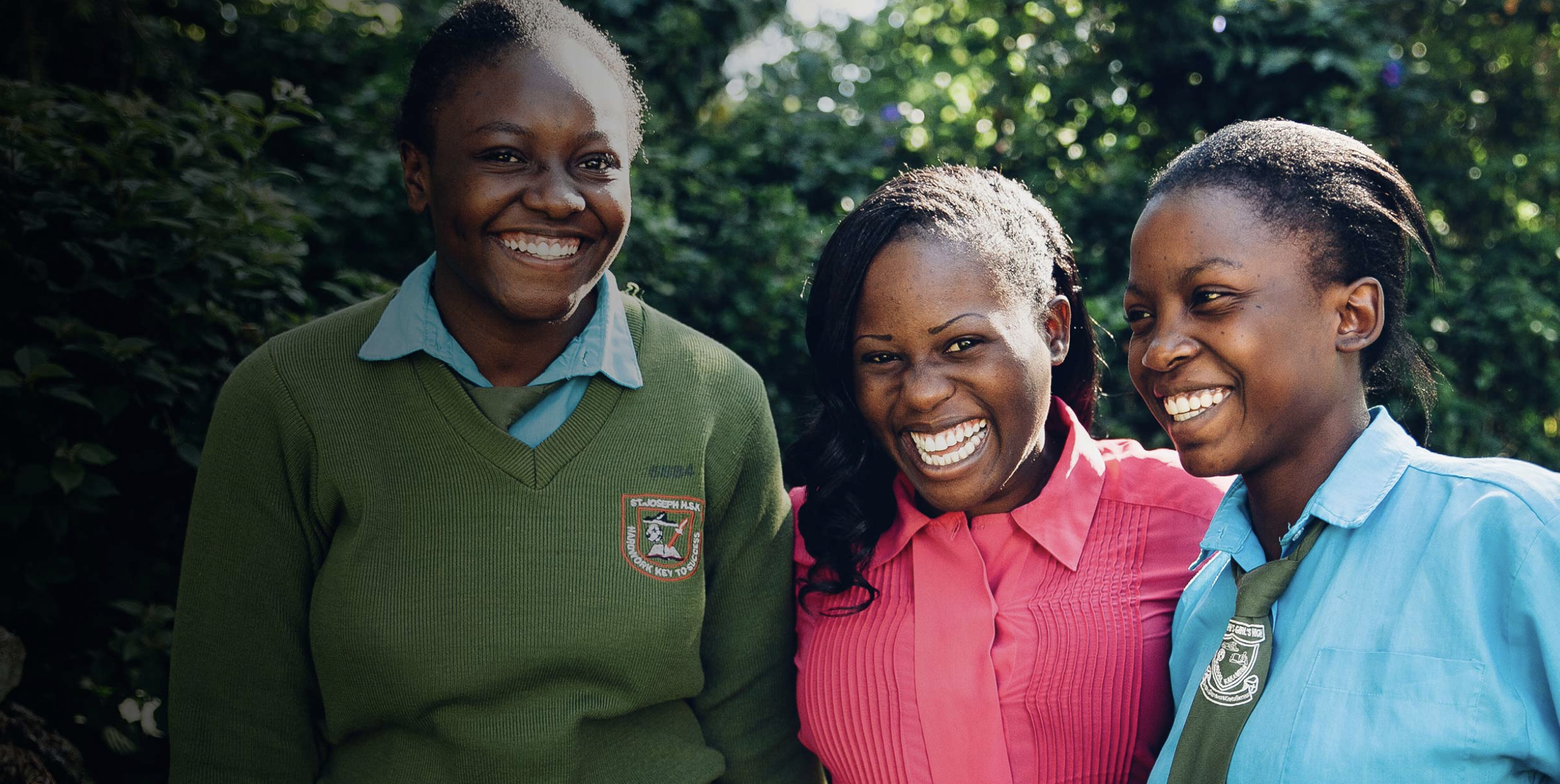 Three people smiling, standing close together outdoors; two in school uniforms, one in a green sweater and another in a blue shirt, with a person in a pink shirt in the middle. Greenery in the background.